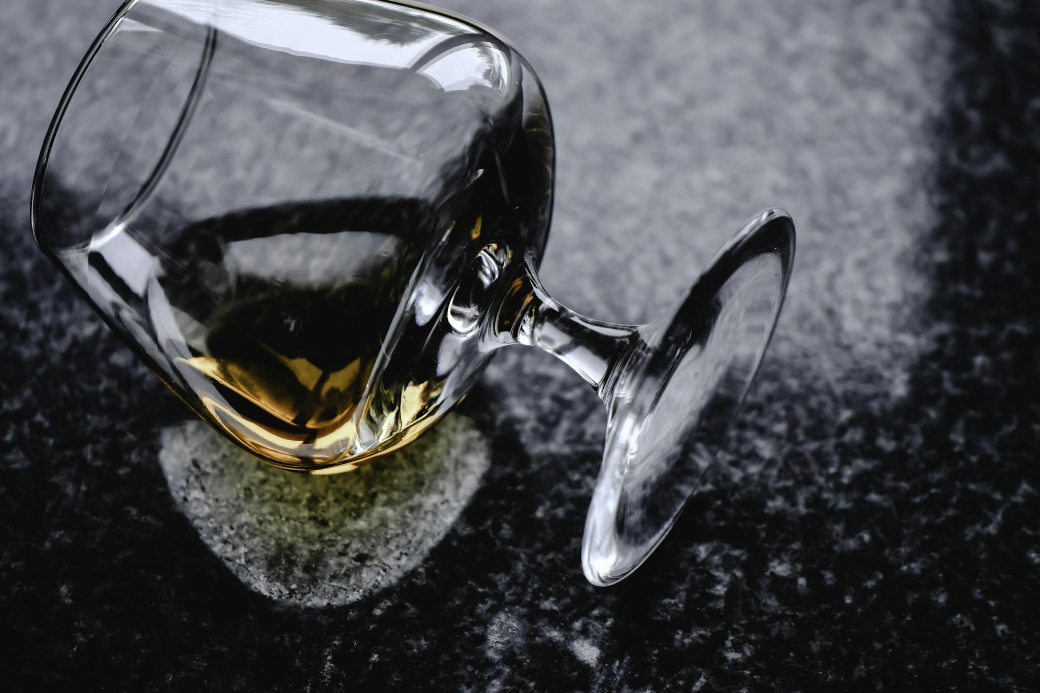 A delicate whiskey glass with liquor spills gracefully on a polished granite surface.