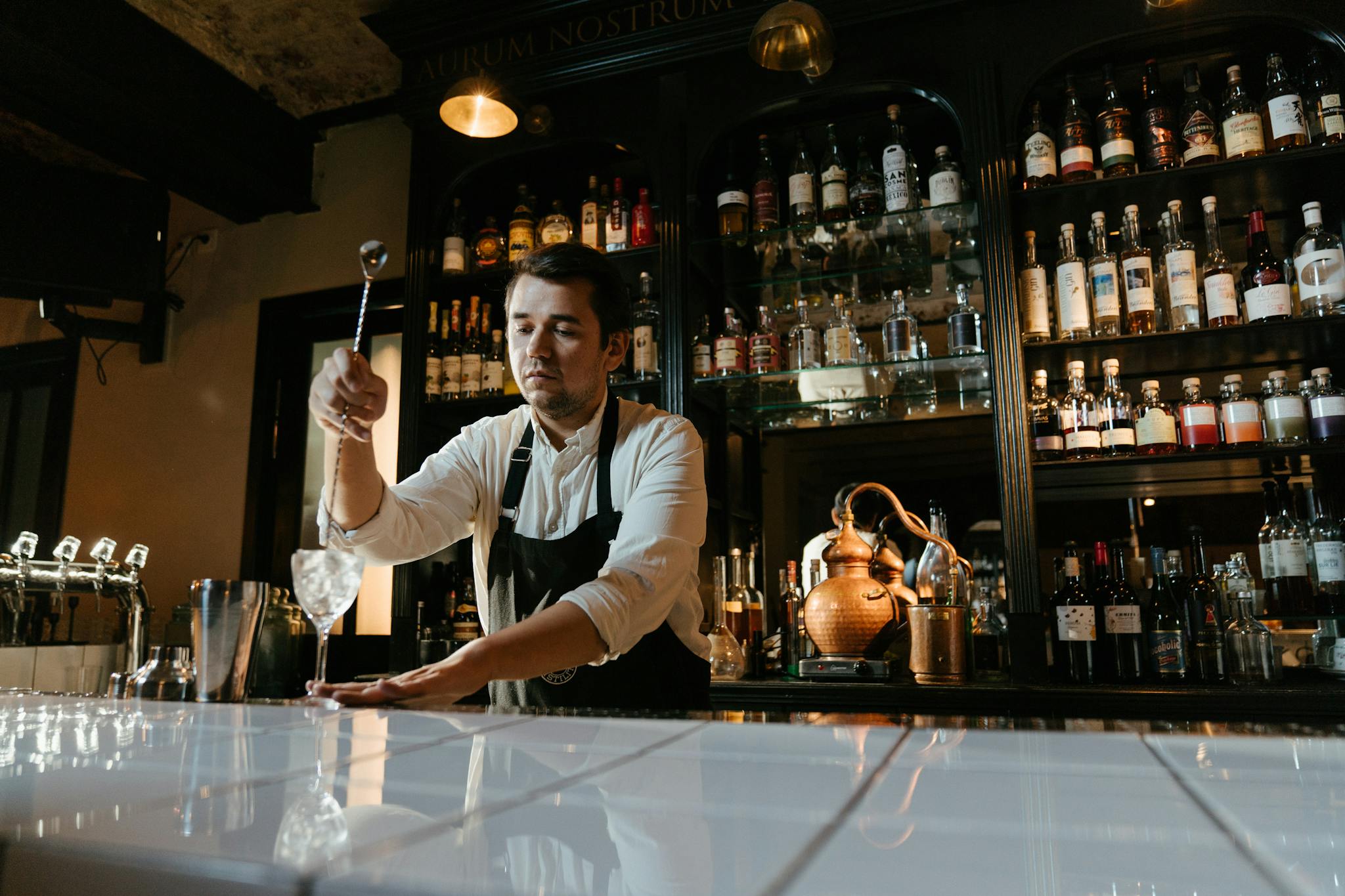 Male bartender mixing a cocktail behind a well-stocked bar. Atmospheric dim lighting indoors.